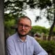 Lähikuva miehestä, jolla on silmälasit ja parta, vaaleansininen paita päällä, puut taustalla.